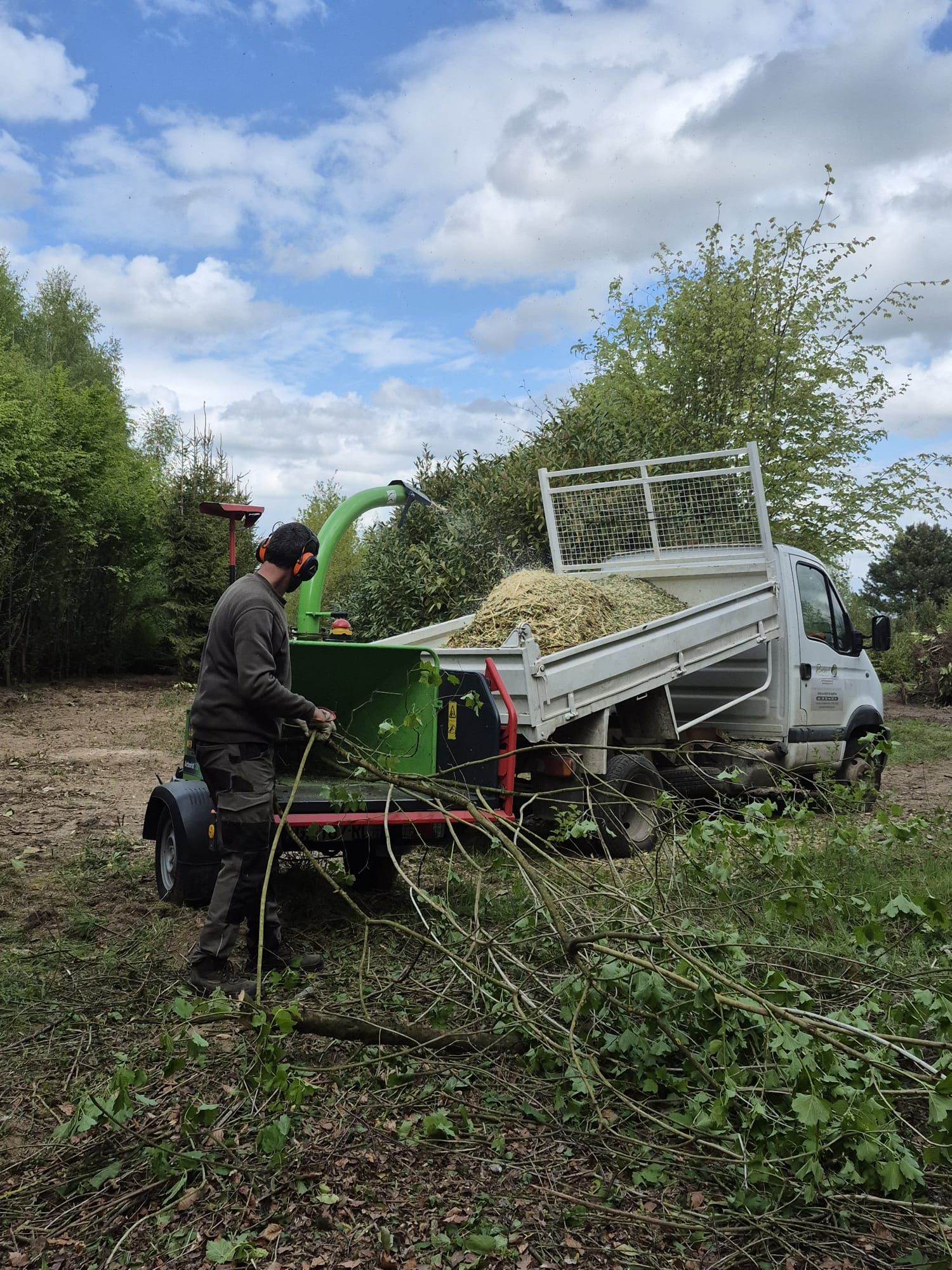 Un ouvrier manœuvrant une déchiqueteuse à bois verte à côté d'un camion blanc dans un champ boueux et envahi par la végétation.