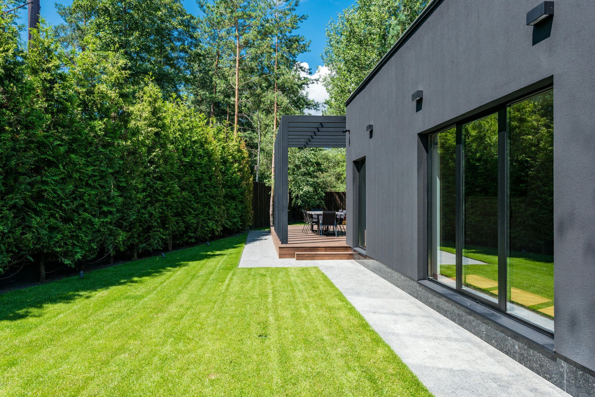 Maison moderne avec pelouse verdoyante, terrasse et grandes fenêtres donnant sur les arbres.