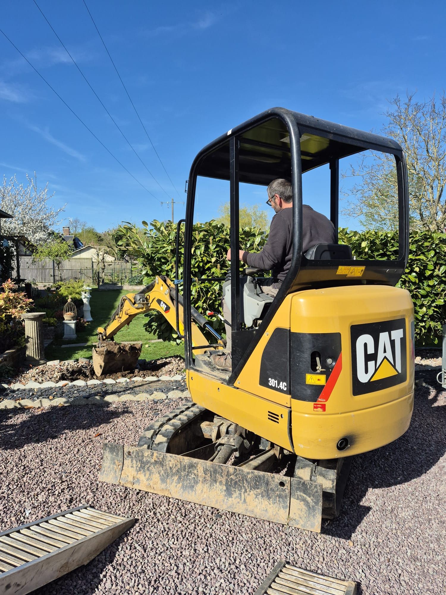 Une mini-pelle CAT jaune sur du gravier avec un opérateur dans une pépinière sous un ciel bleu clair