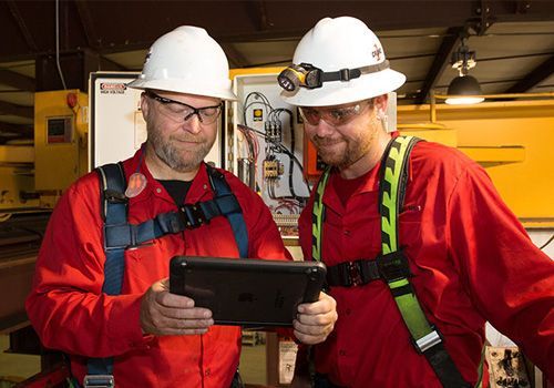 Two workers in red coveralls and helmets, looking at a tablet in a machinery room.
