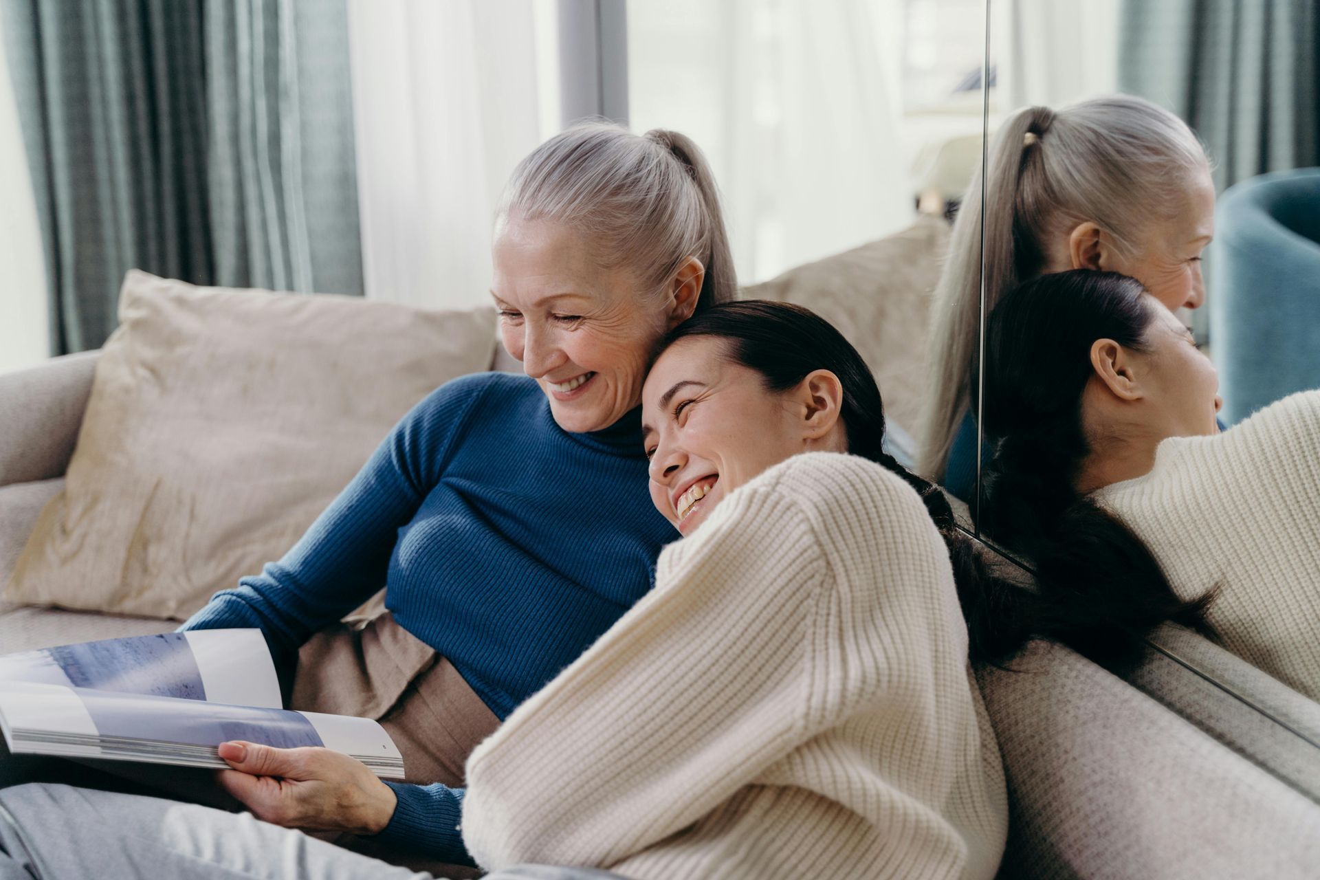 Older woman and young woman on a sofa, reading a book and smiling. A mirror reflects their images.