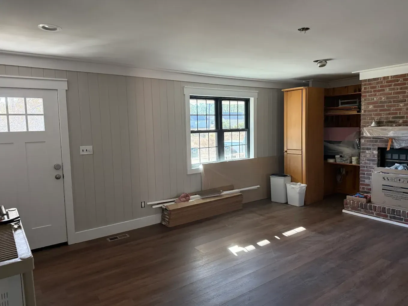 An empty living room with wood flooring, painted paneling, a brick fireplace, and a front door.