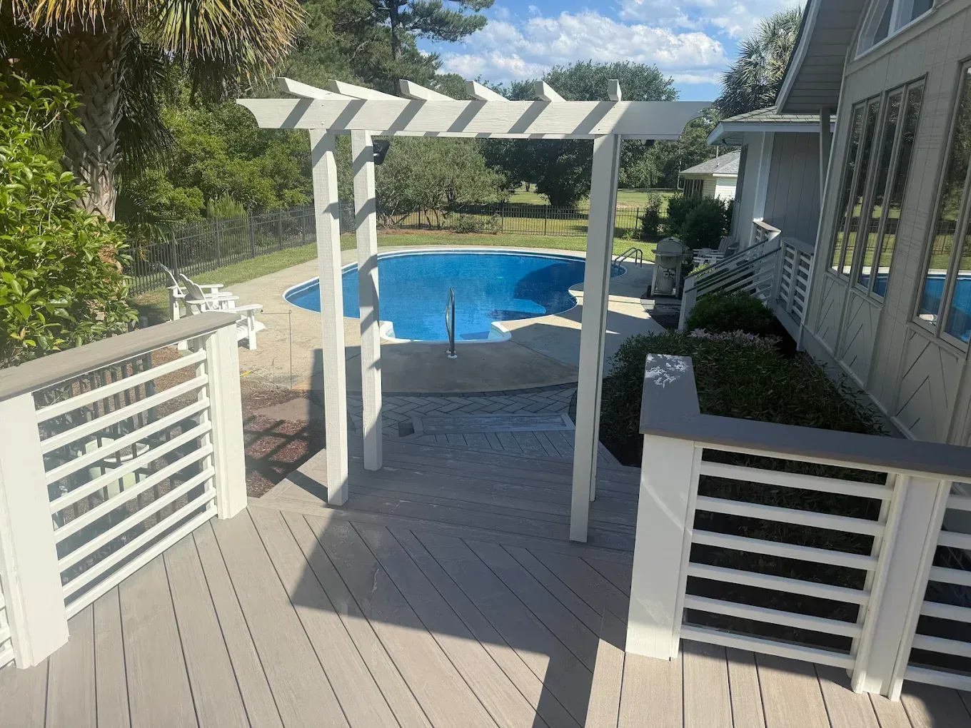 Deck with a white pergola, overlooking a blue pool and green trees.