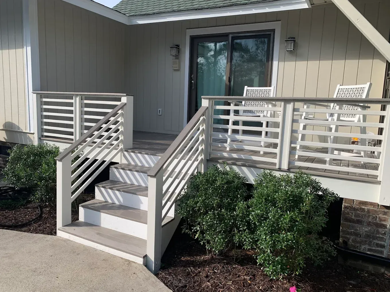 A small deck with steps leading to a sliding glass door. Beige railing, white chairs, and green bushes.