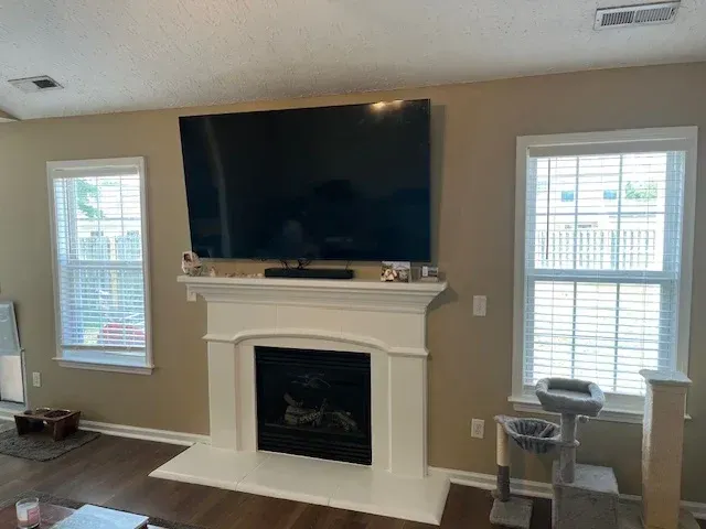 A living room with a fireplace and a large TV mounted above it, flanked by two windows.