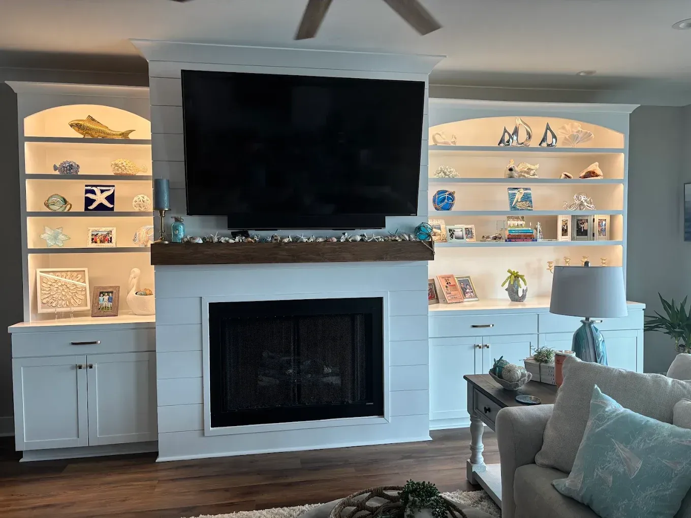 White built-in cabinets flank a fireplace with TV. Shelves display coastal decor. A lamp sits on a side table.