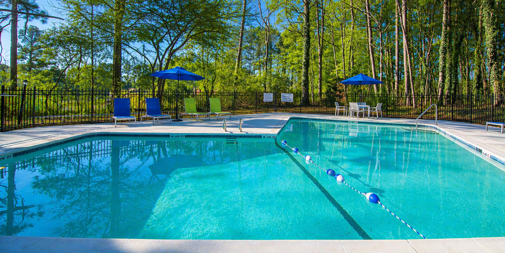 A clear, turquoise swimming pool with blue umbrellas, chairs, and a treeline in the background on a sunny day.