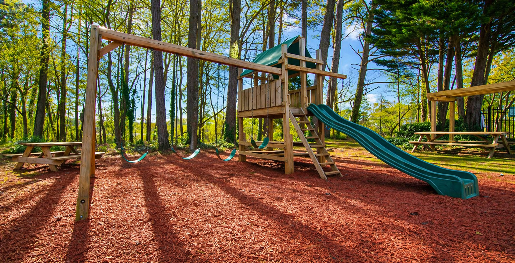 Playground in a wooded area with swings, slide, and wooden structure; ground covered in wood chips.