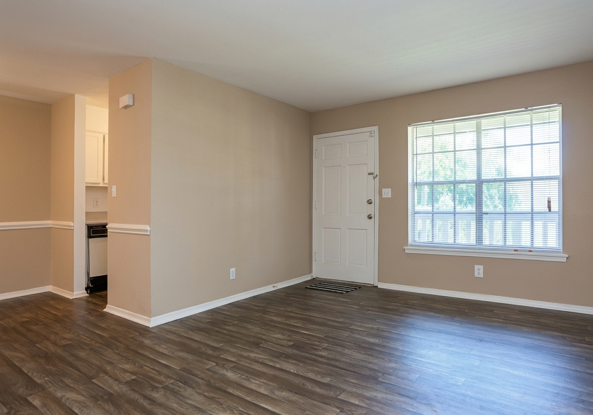Empty living room with wood-look flooring, beige walls, and a window; door leads to a kitchen.