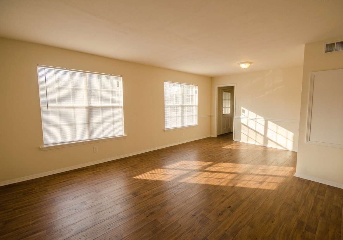 Empty room with wood-look flooring, beige walls, two windows with blinds, and a doorway.