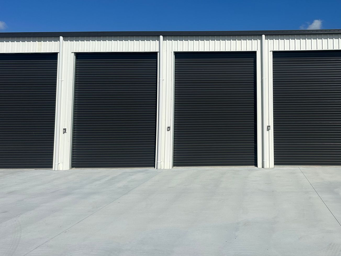 Row of dark storage unit doors beneath a blue sky, with a concrete driveway in front