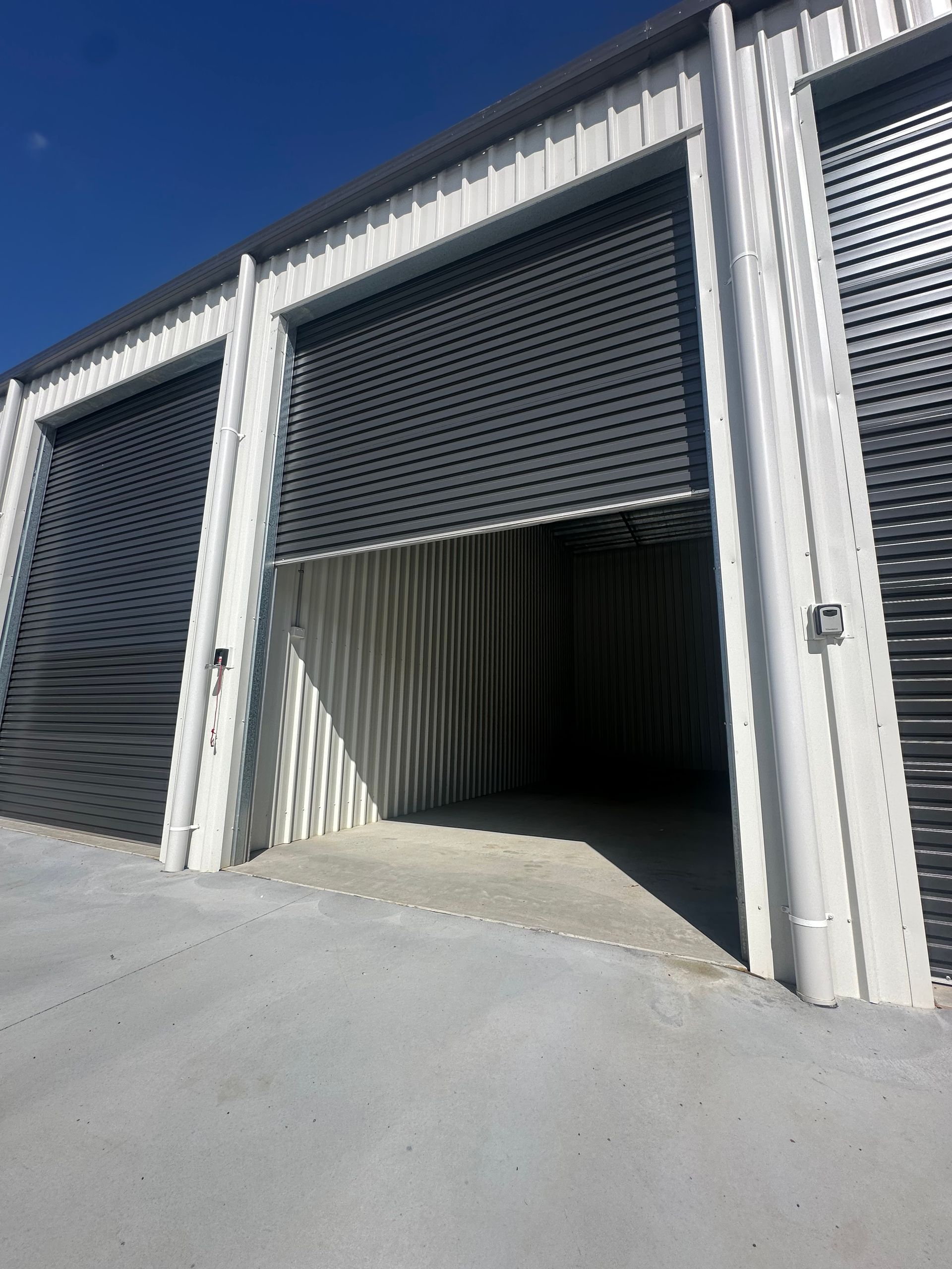 Industrial Storage Units With Metal Roll-up Doors, One Partially Open To A Dark Interior Under A Blue Sky — Midcoast Premium Caravan Storage in Mid North Coast, NSW