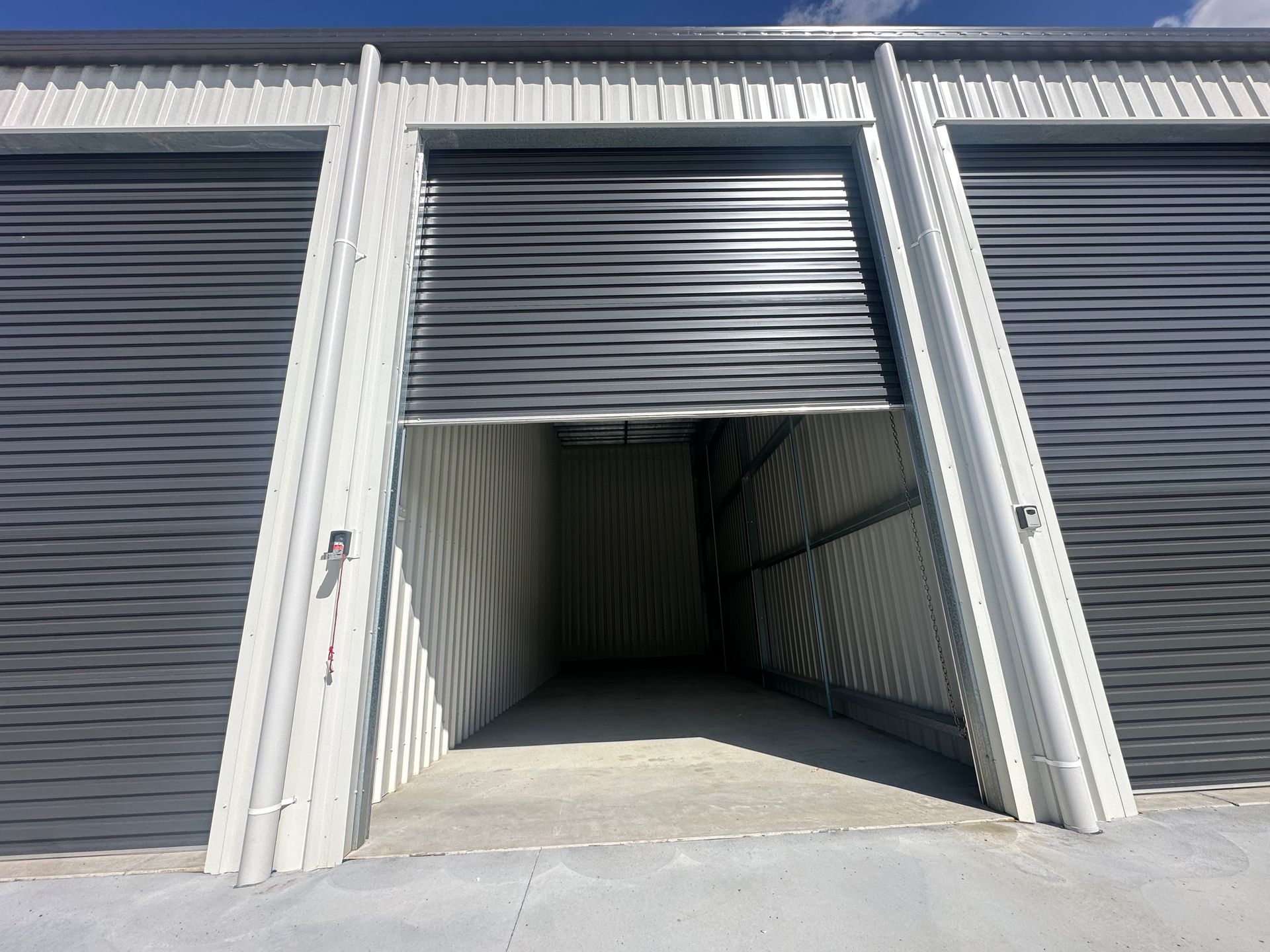 Open storage unit with raised metal roll-up door and dark interior between closed gray doors — Midcoast Premium Caravan Storage in Forster, NSW