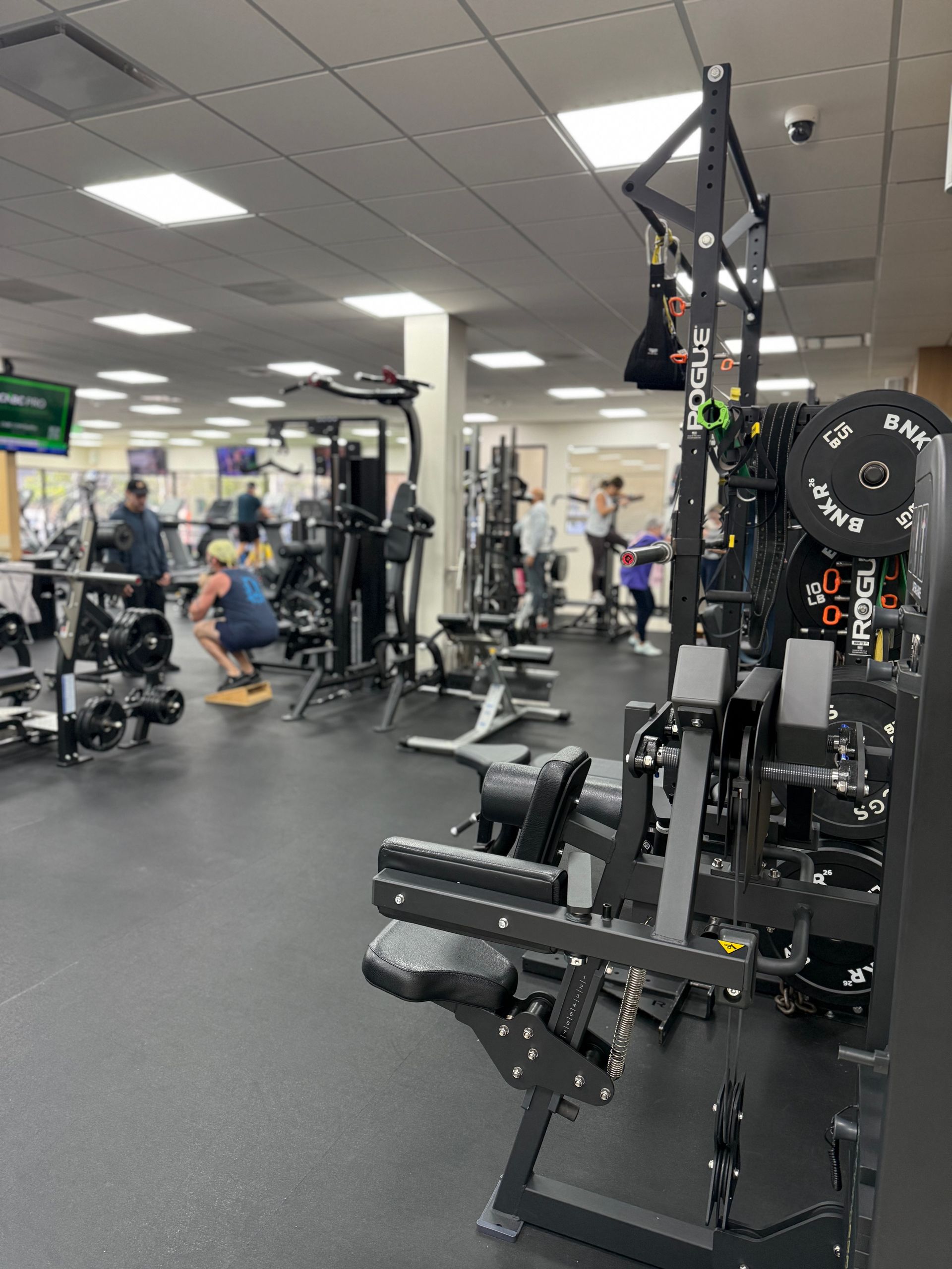 Gym interior with various exercise machines and people working out. Black floor, bright lighting, and equipment.