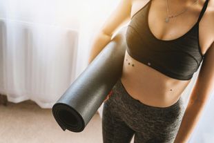 Woman holding a rolled-up yoga mat, wearing a sports bra and leggings, by a window.