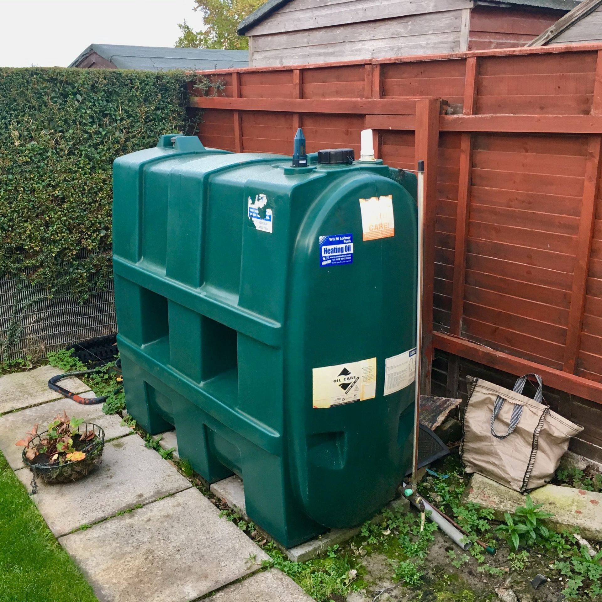 Photograph of a Harlequin 1100SL Slimline Heating Oil Tank, atop a concrete base, with fence and red fence in the background.
