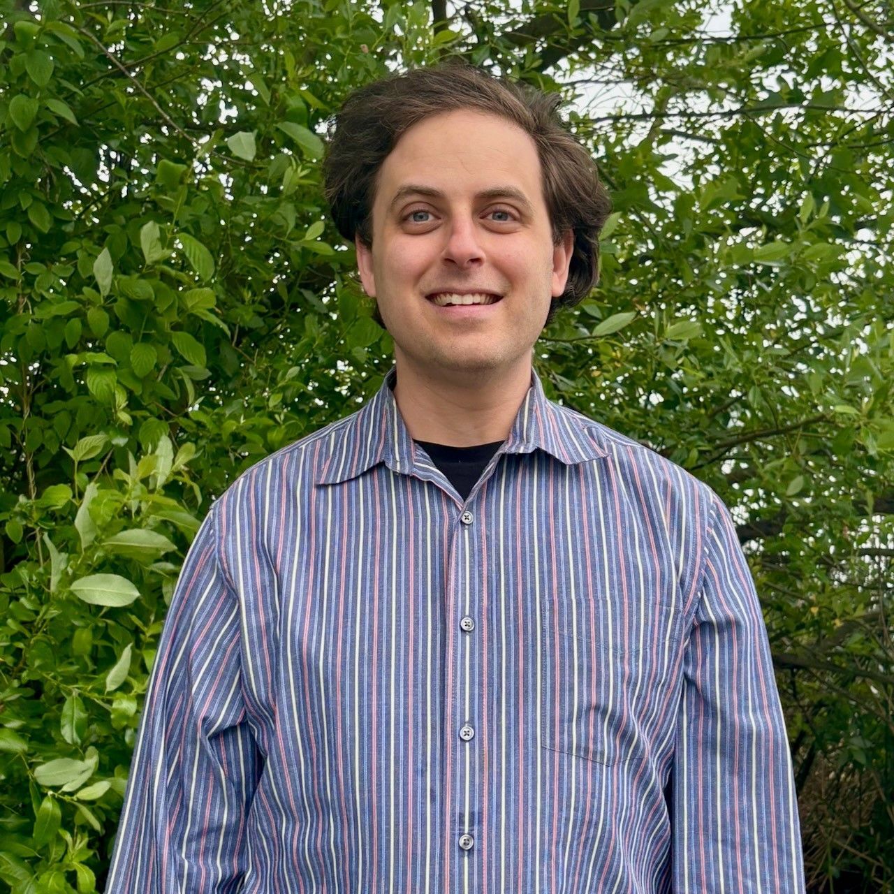 A man in a striped shirt is standing in front of trees.
