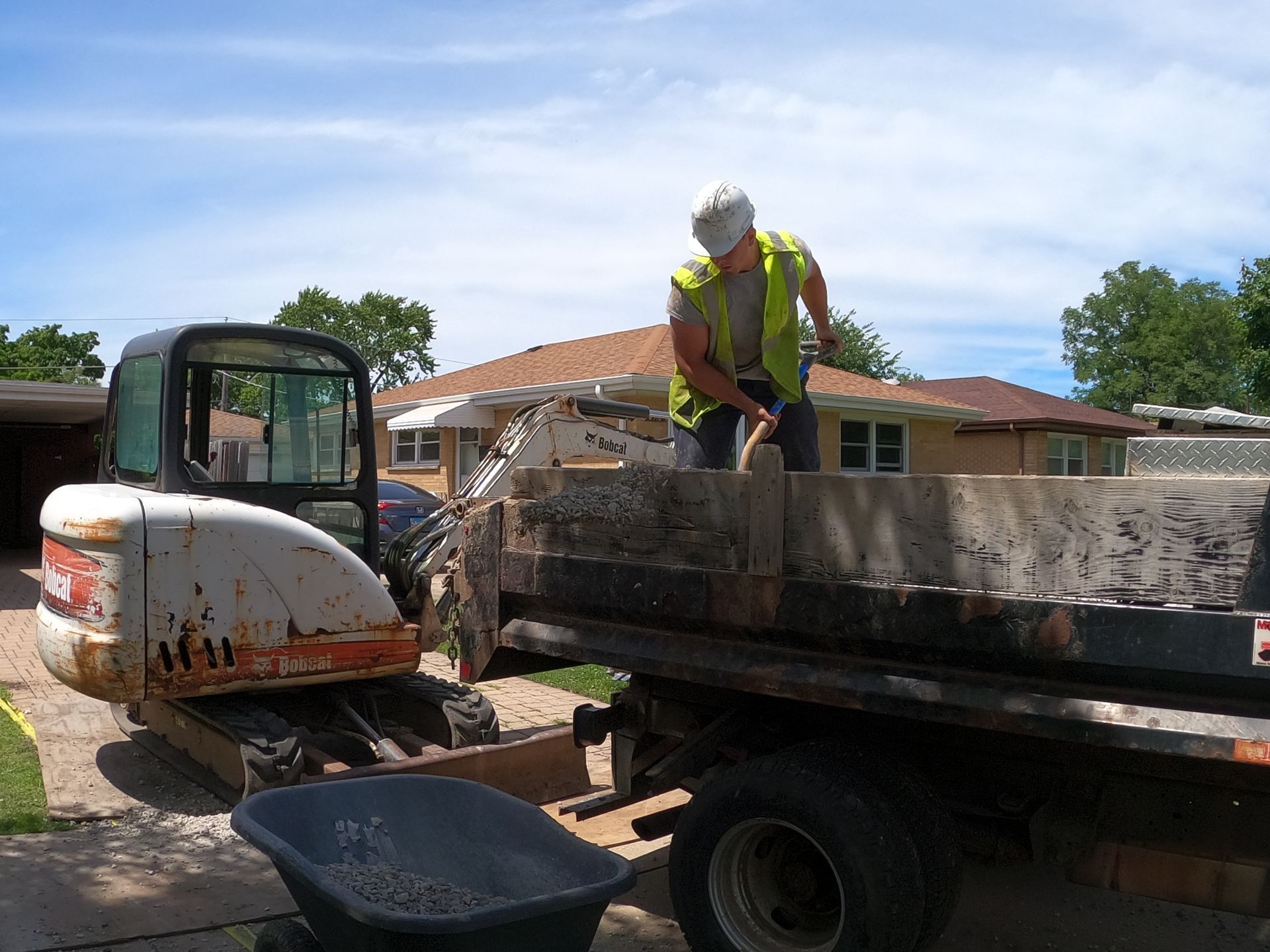 Man in safety vest shovels debris from a truck bed next to a small excavator in a residential driveway.