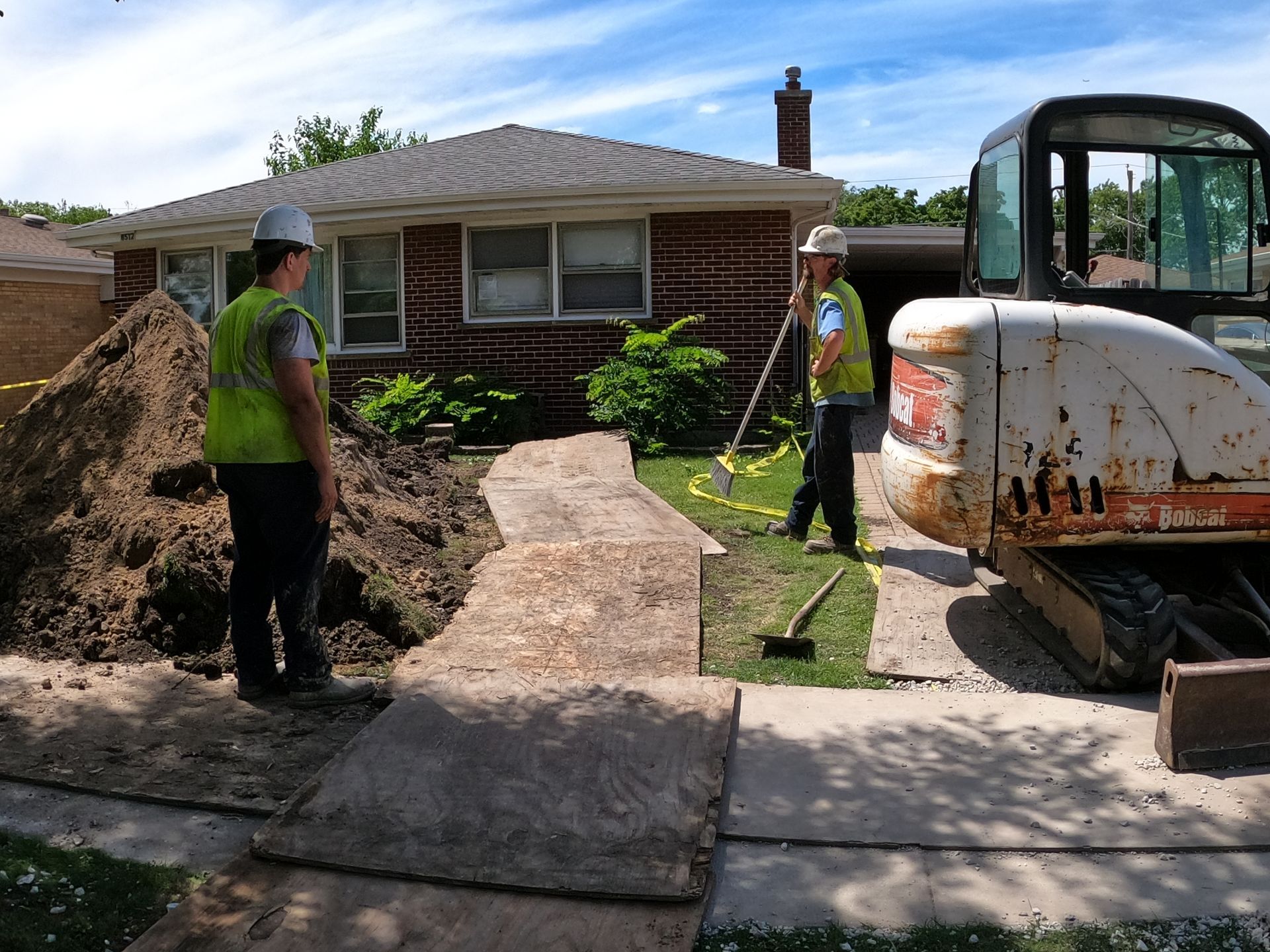 Two workers in safety vests near an excavator and a residential house, with excavated earth and a walkway.