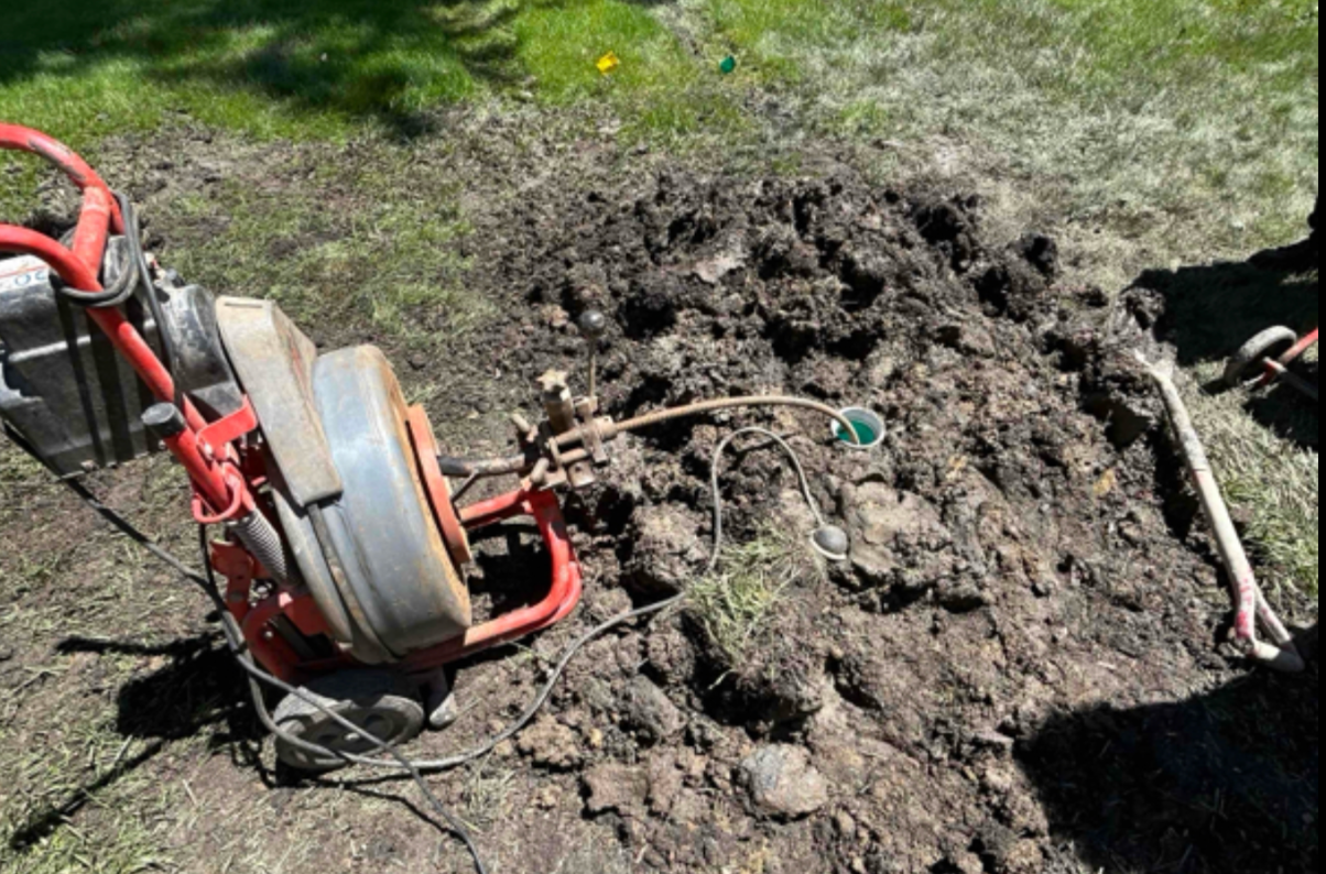 Red sewer snake machine next to a hole dug in the ground, with dirt pile; green cap visible.