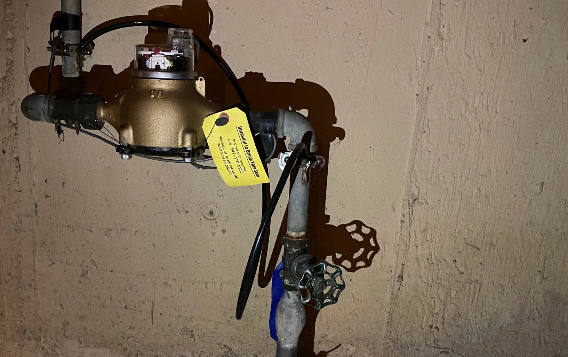 Water meter with pipes and valve against a light-colored wall. A yellow tag hangs from it.