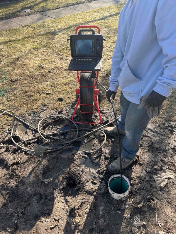 Person using a sewer camera to inspect a pipe; equipment set up on muddy ground.