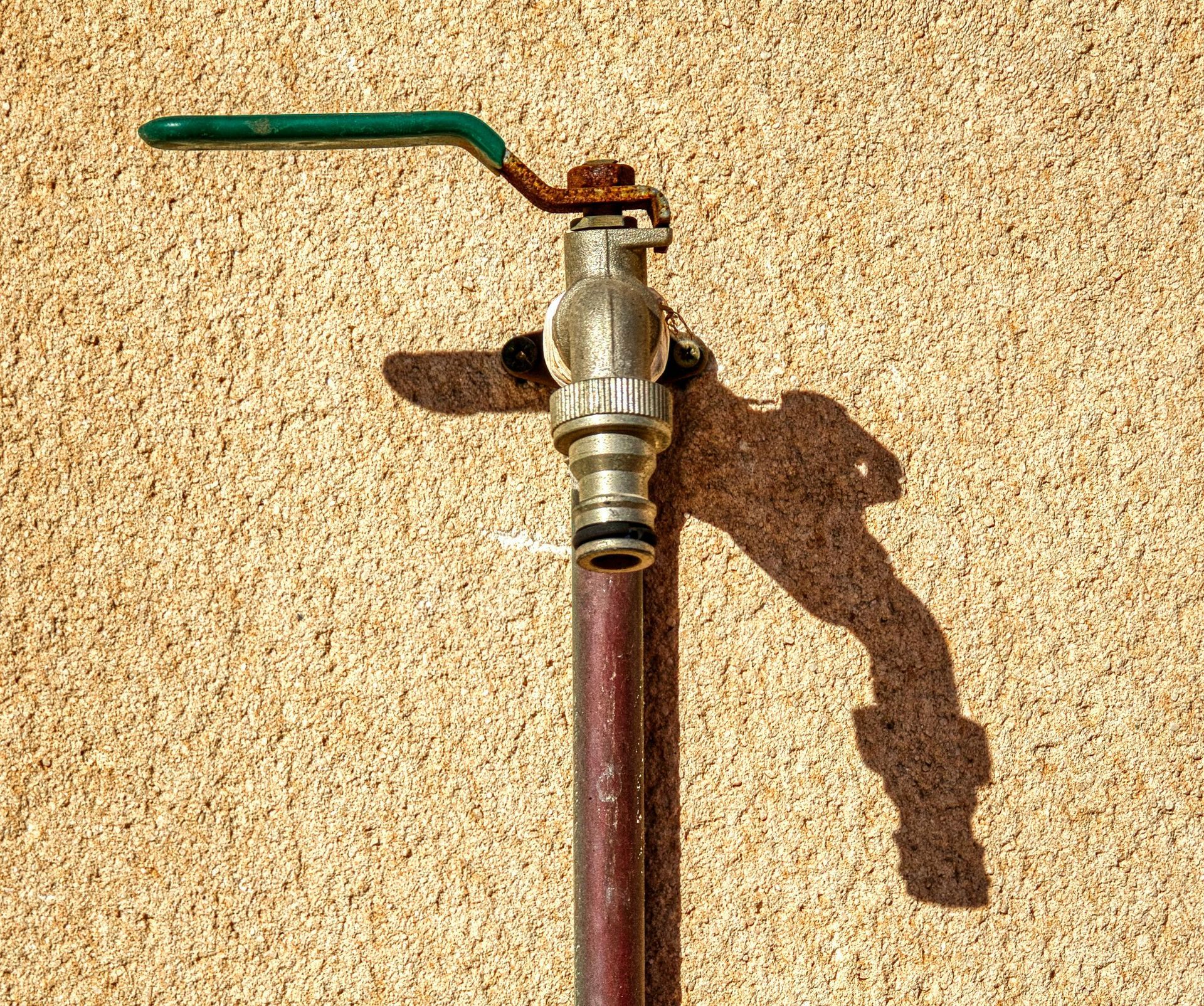 A rusty outdoor water faucet with a green handle on a textured beige wall.
