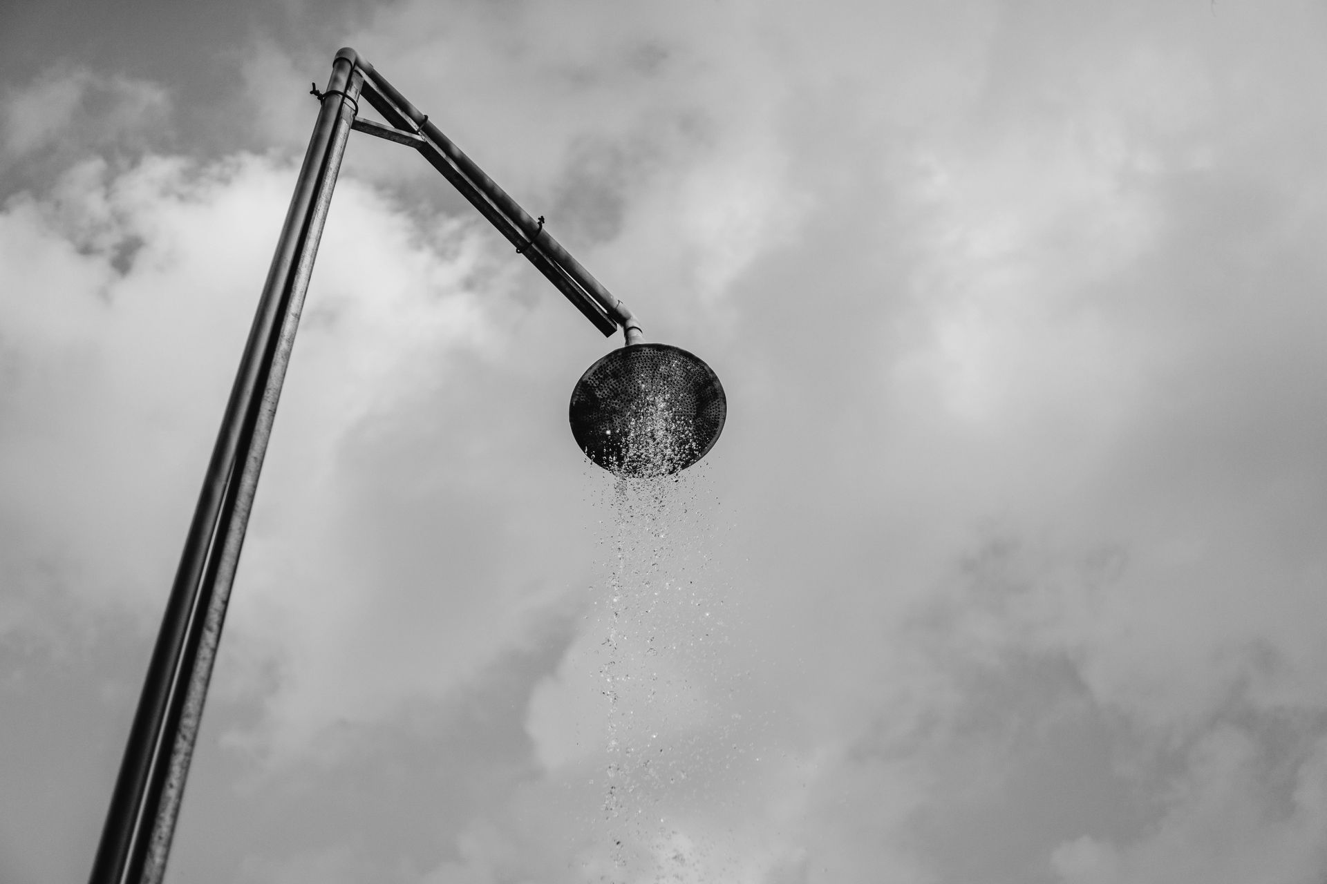 Outdoor shower head with water falling against a cloudy sky.
