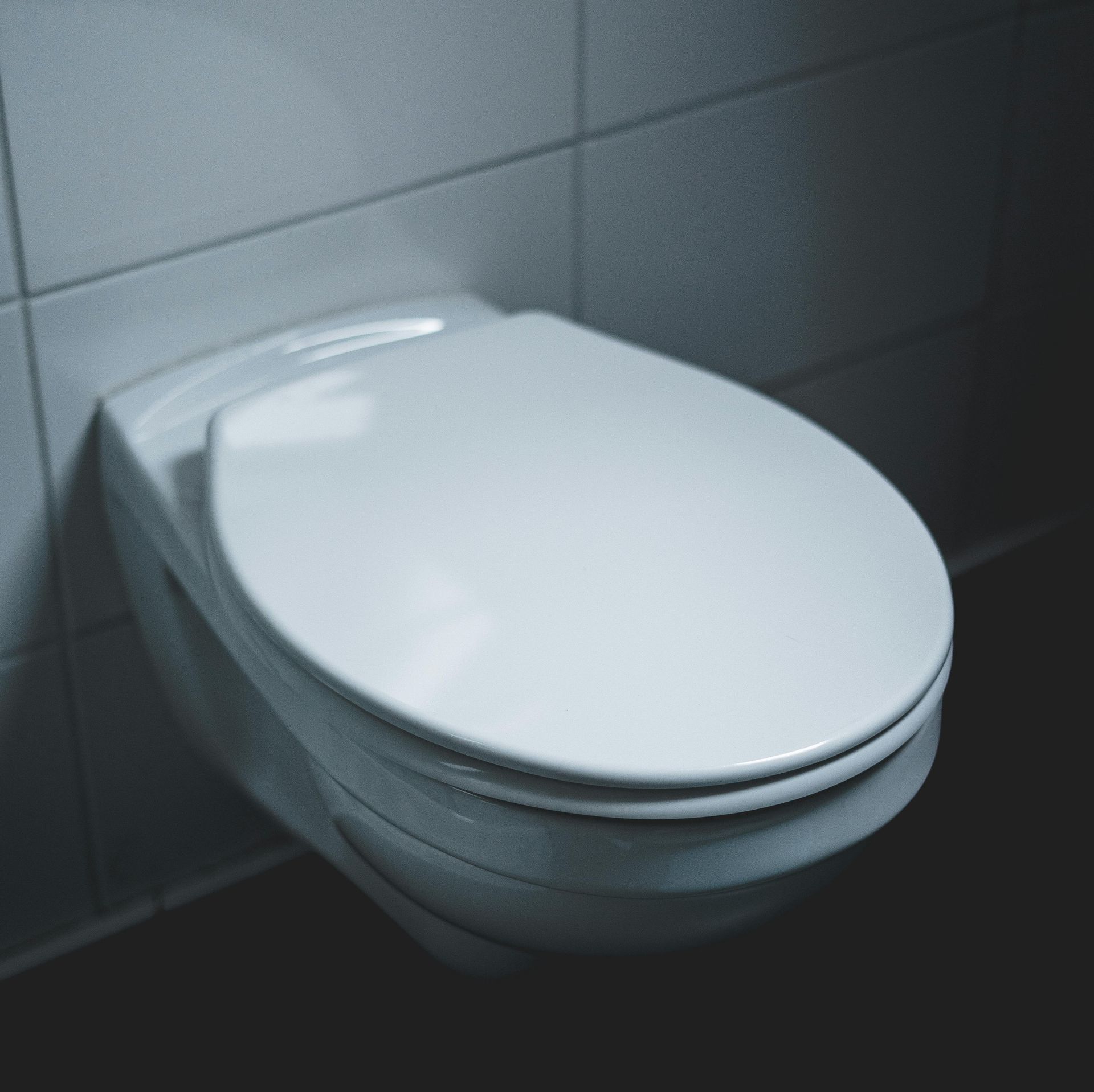 White, wall-mounted toilet with closed lid, against white tiled wall and black floor.