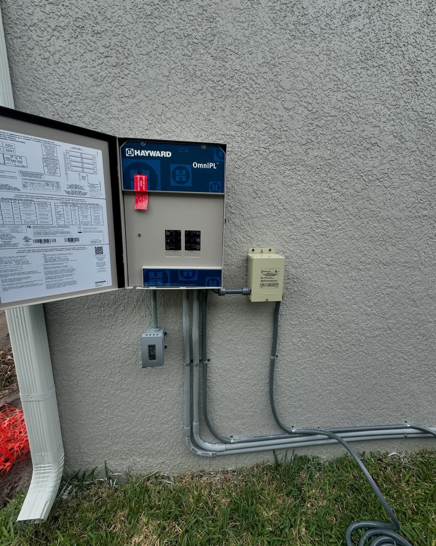 Outdoor electrical control panel mounted on a wall beside a sign and conduit pipes
