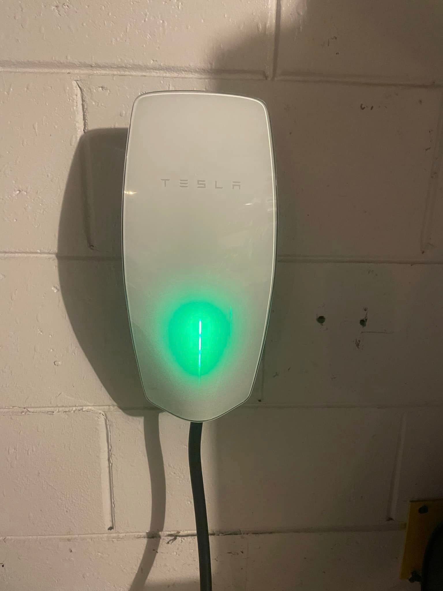 White wall-mounted EV charger with a glowing green light on a cinderblock wall.