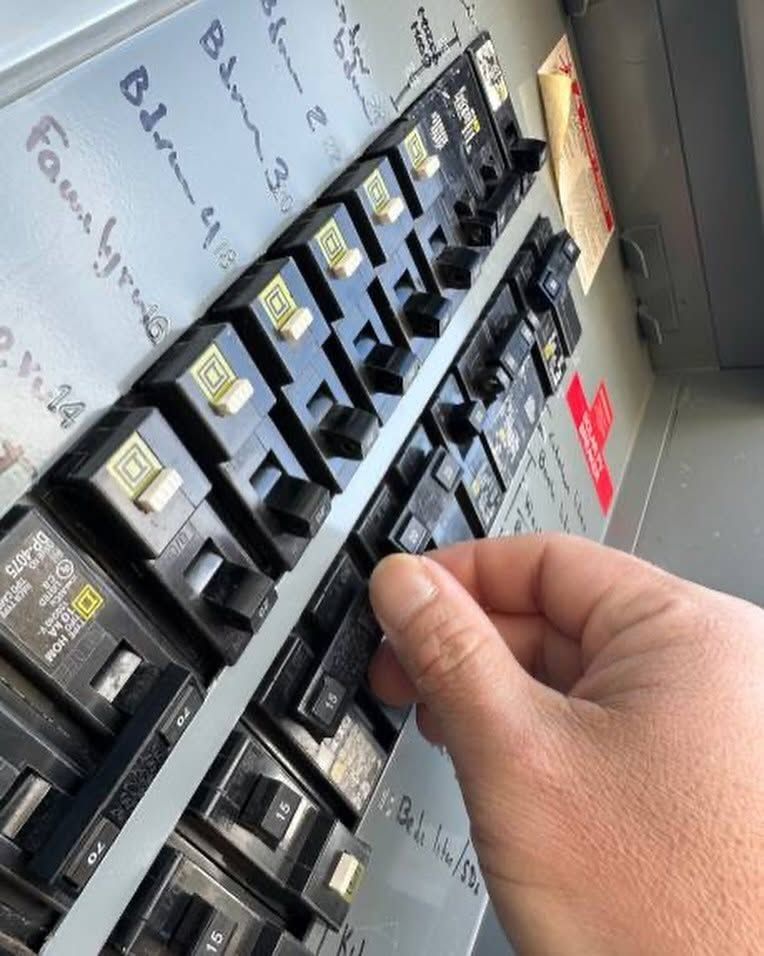 Hand turning a circuit breaker in an electrical panel with labeled switches and red warning tags.