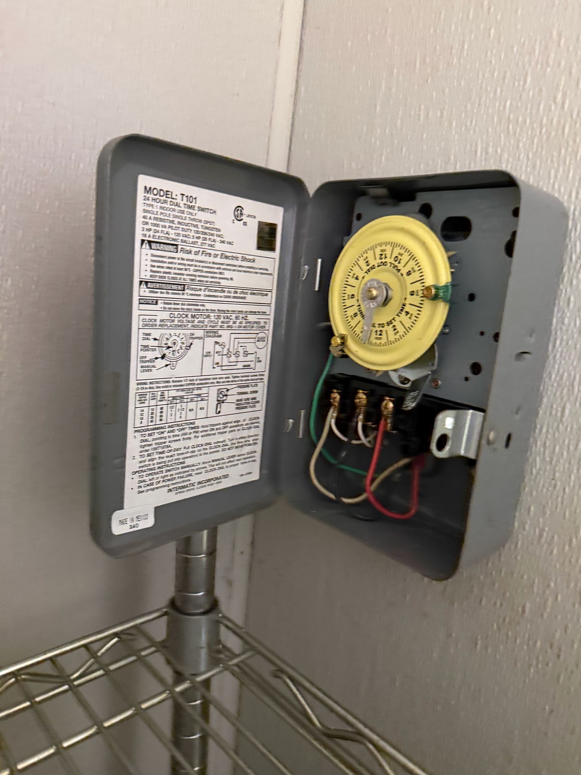 Open electrical control box with yellow timer mounted on a wall above a metal shelf.