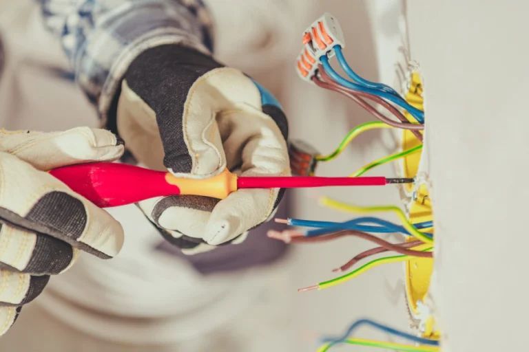 Gloved hands use a red screwdriver on exposed multicolored electrical wires in a wall outlet box