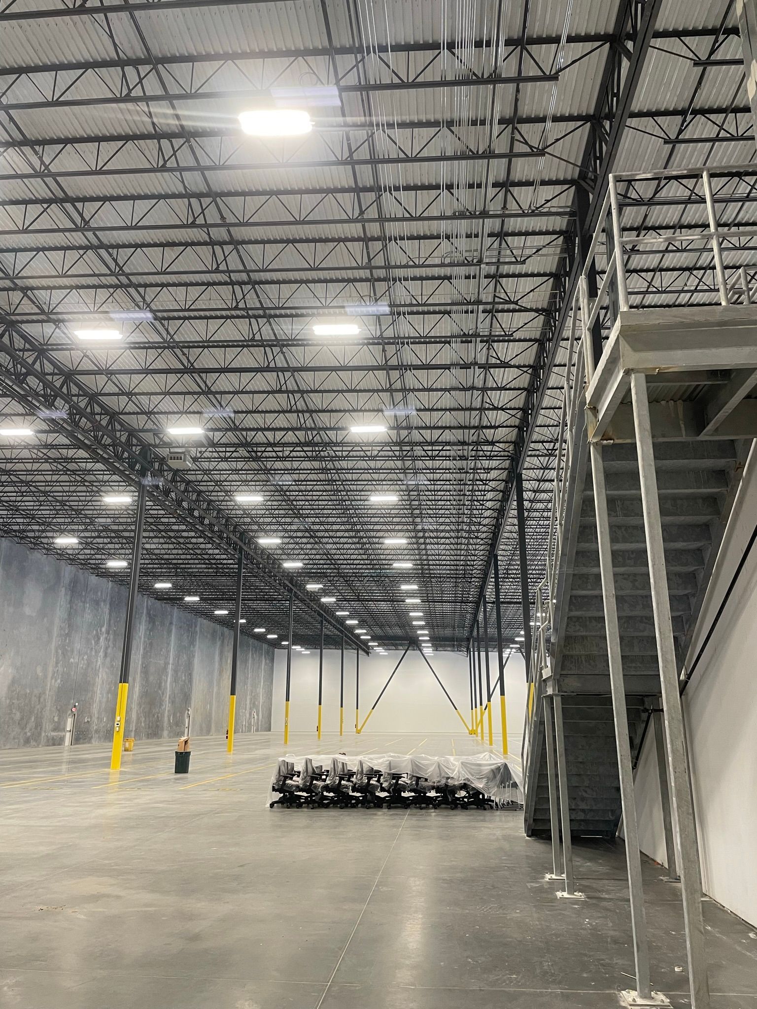 Large empty warehouse interior with high metal ceiling, bright overhead lights, and stacked pallets near a stairway