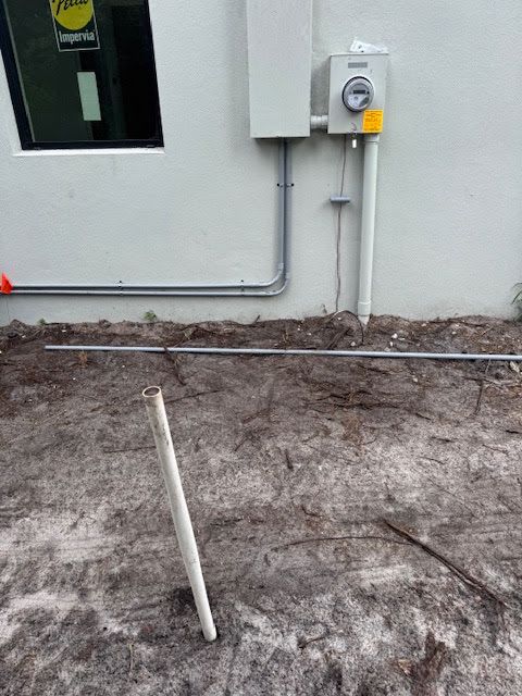 Outdoor utility area with wall-mounted electrical box, conduit pipes, and disturbed dirt ground with a white stake.