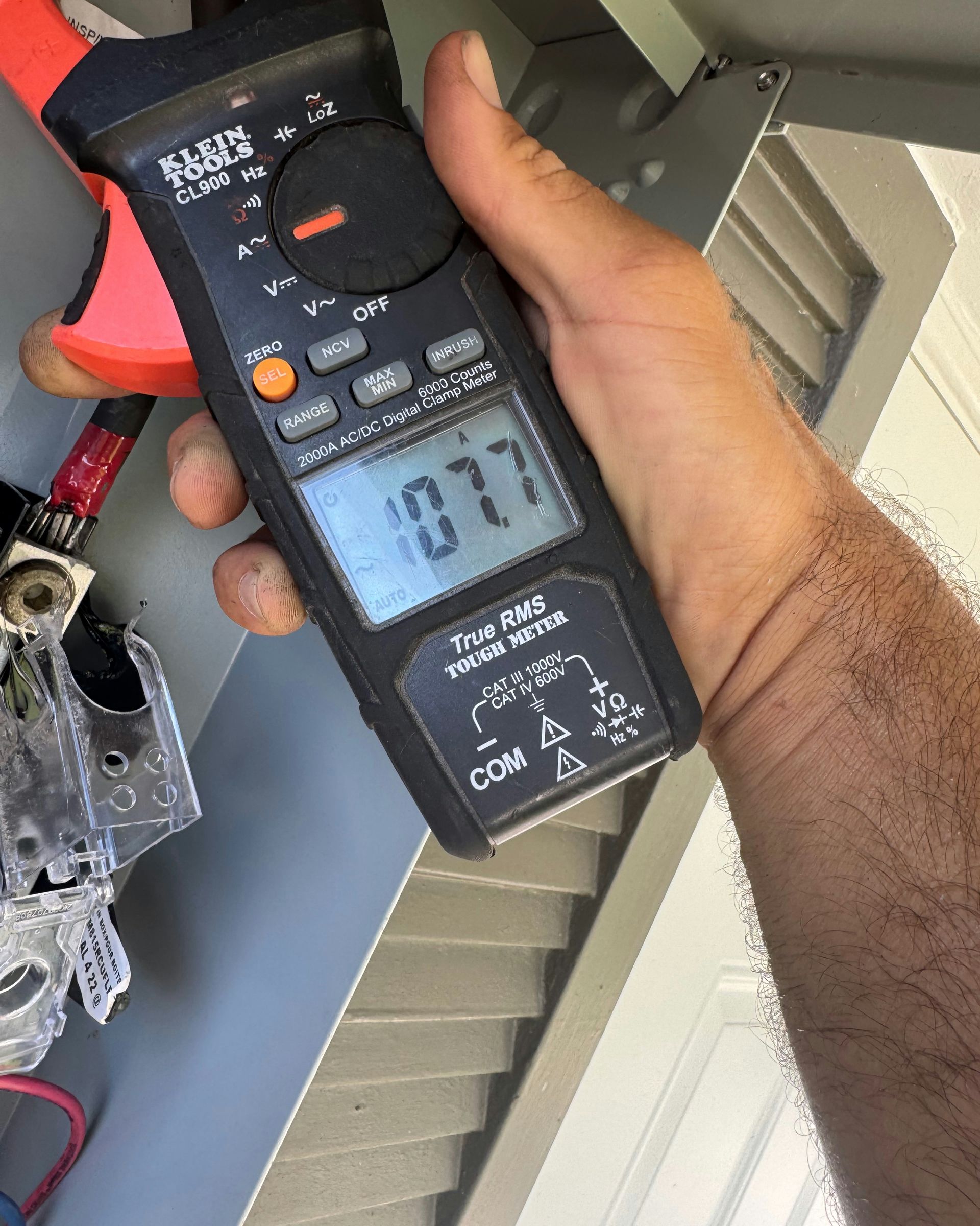 Hand holding a digital multimeter with a 10.7 reading, testing a wire near a vented panel.
