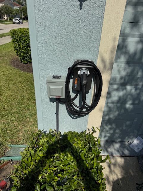 Outdoor EV charger mounted on a house exterior beside a hedge and sidewalk.