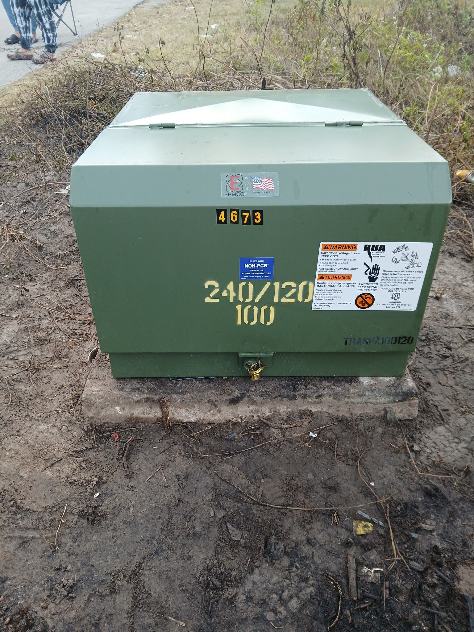 Green utility transformer box outdoors on dirt, labeled 24V/120V, with warning stickers.