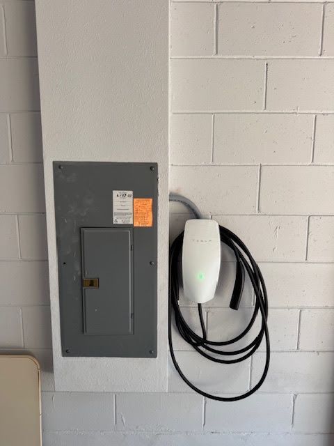 Gray electrical panel with an EV charging unit and coiled black cable mounted on a white cinderblock wall