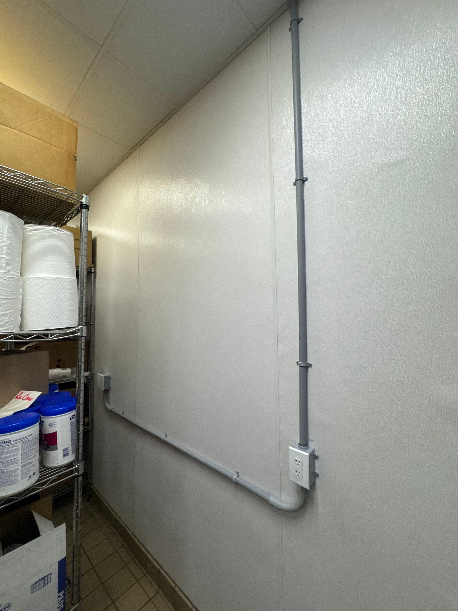 Narrow storage room with metal shelving, cleaning supplies, and conduit running along the wall.
