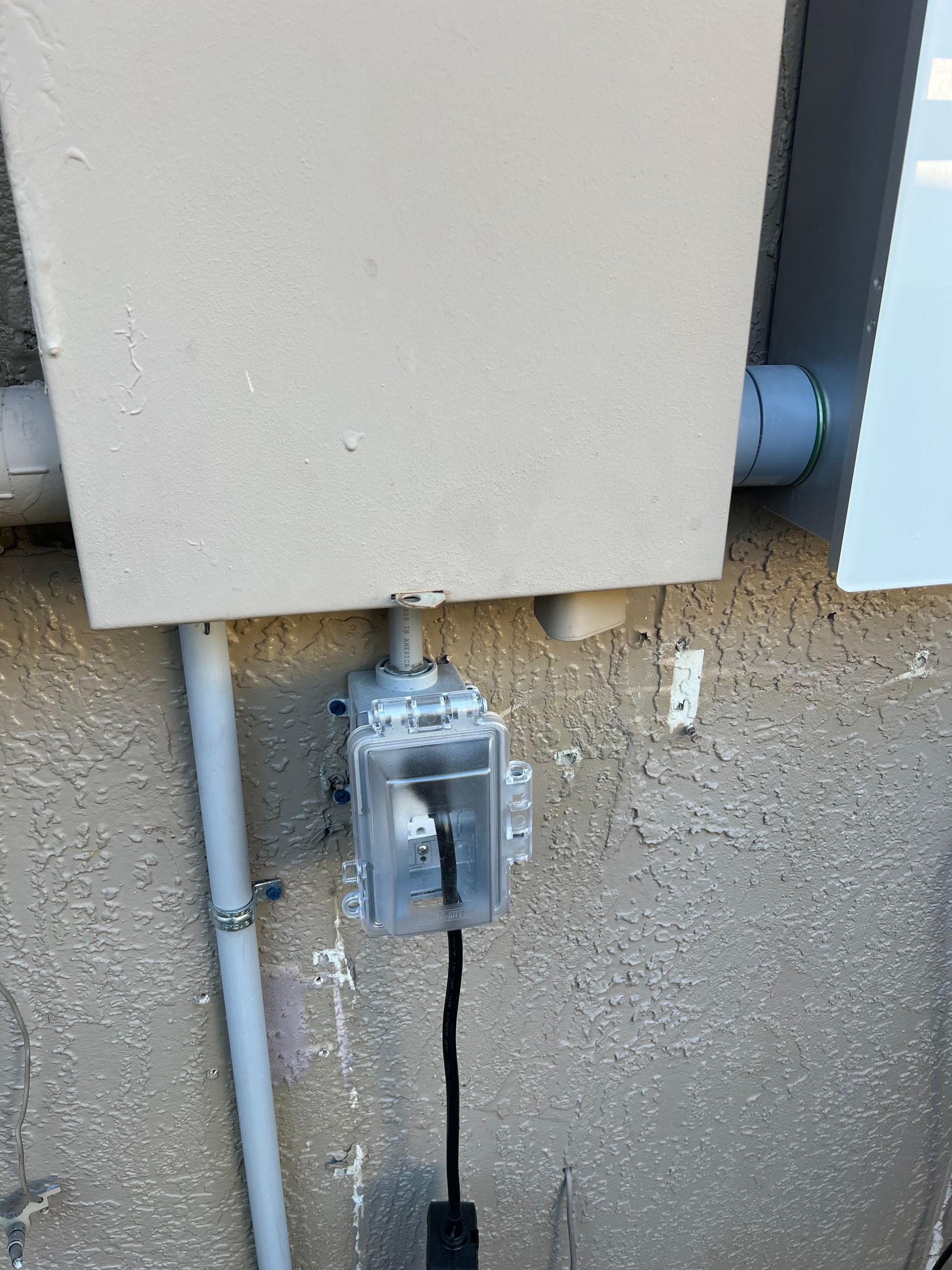 Outdoor electrical box with transparent cover mounted on a stucco wall, with conduit and a black cord below.