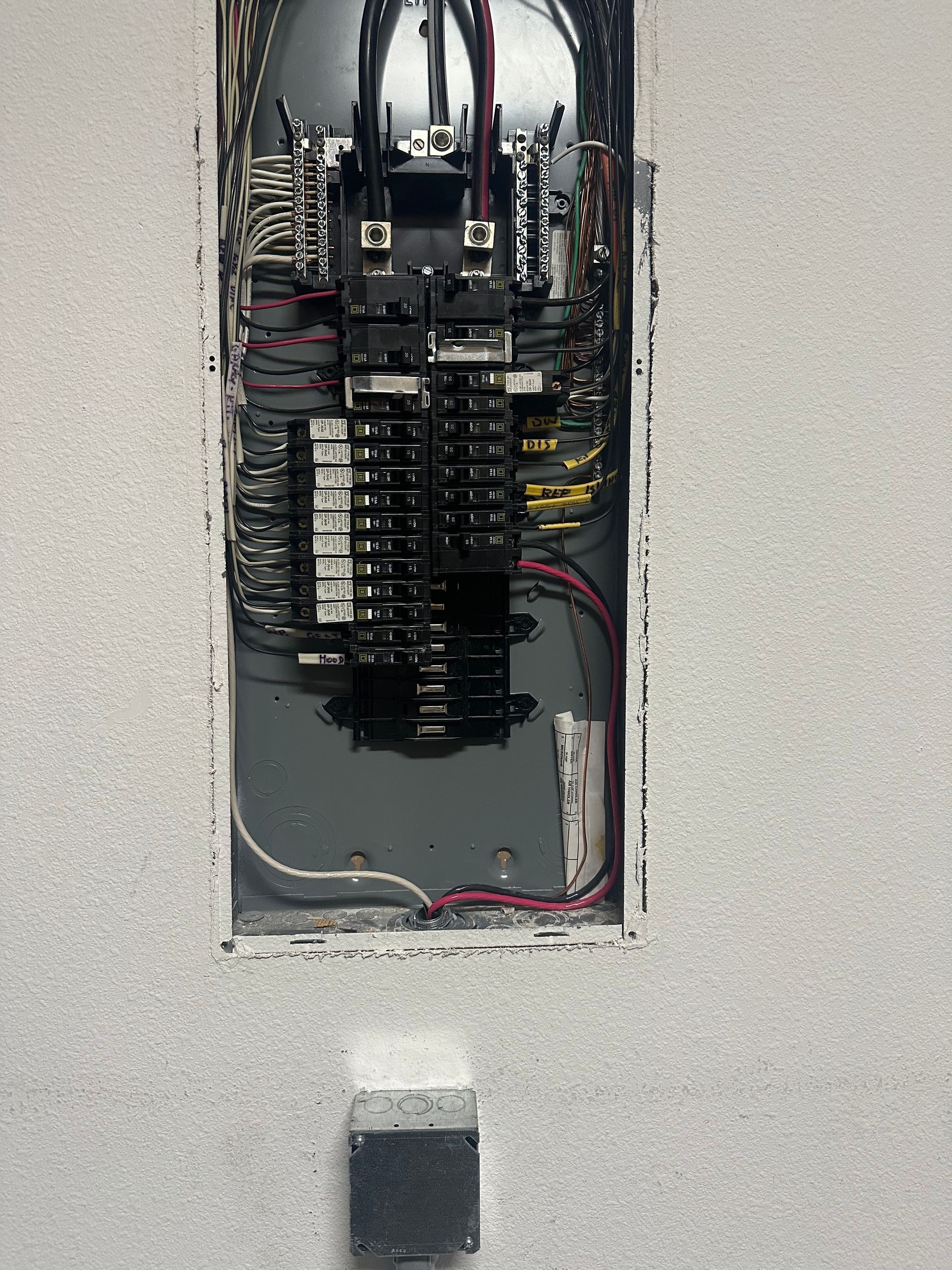 Open electrical breaker panel in a wall with exposed wiring and breakers.