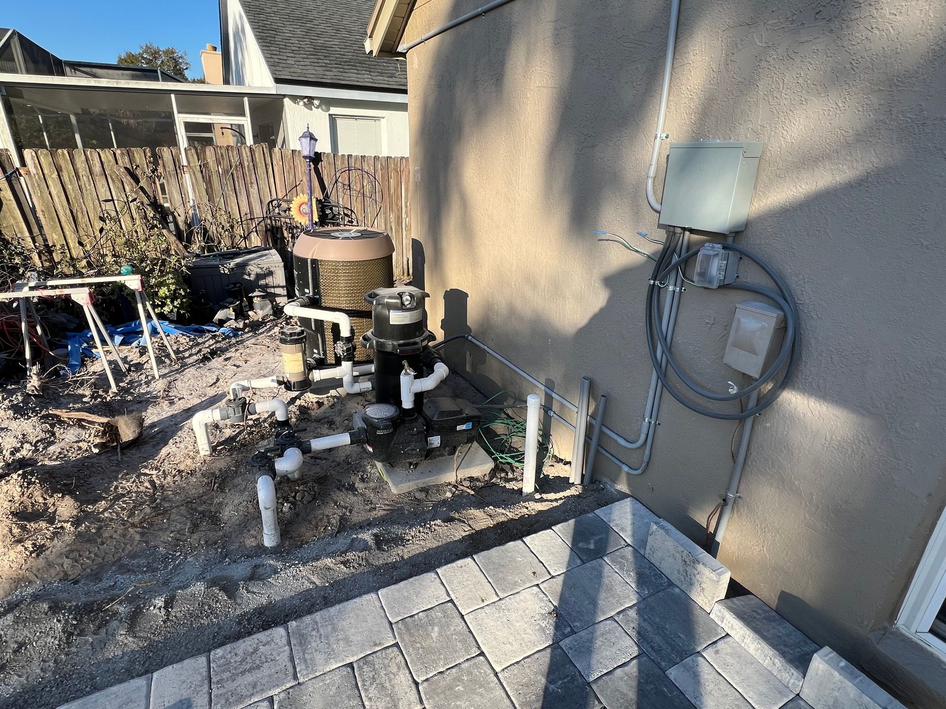 Backyard patio with exposed pipes, utility equipment, and a hose on a beige stucco wall.