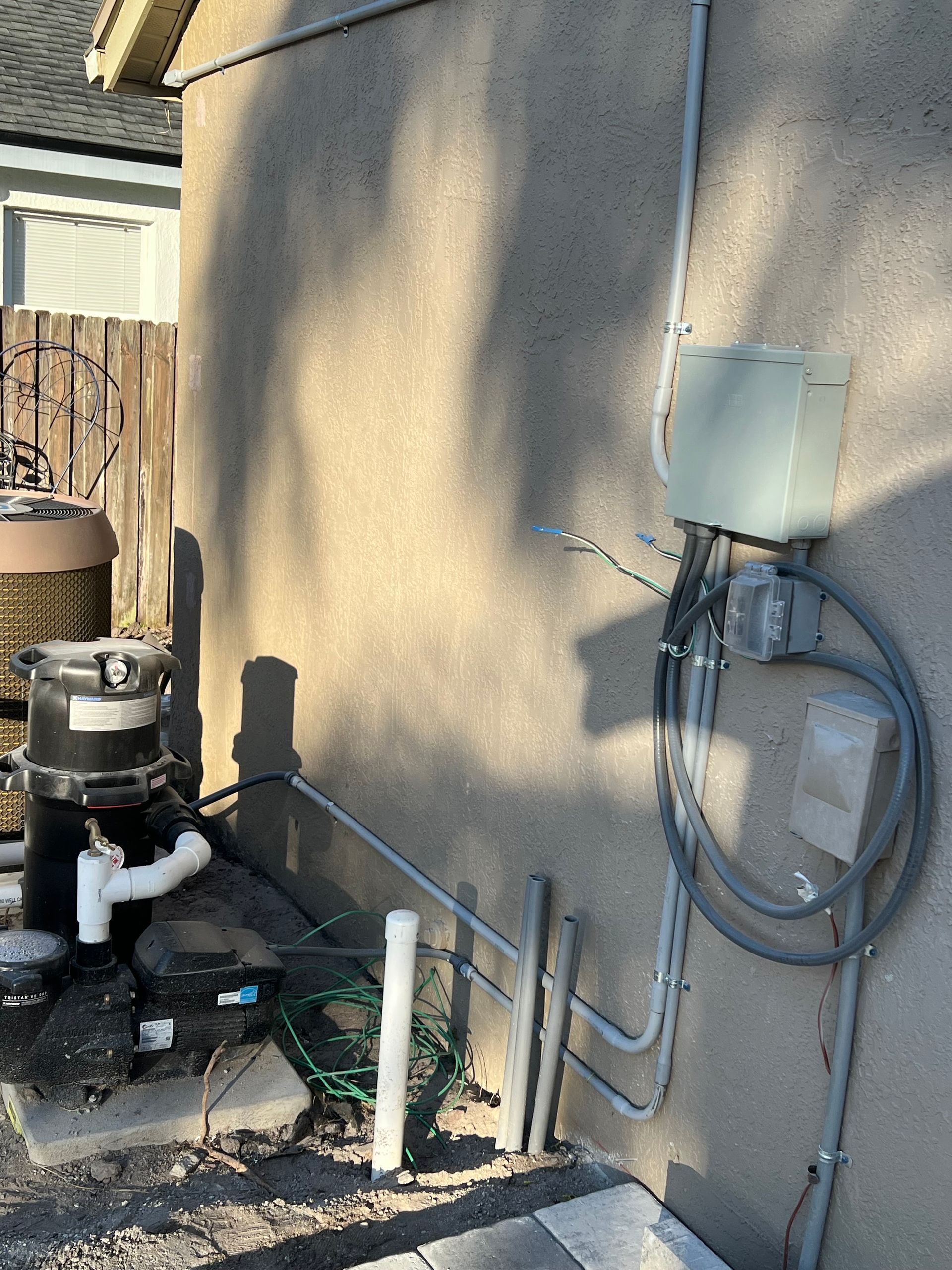 Outdoor utility equipment and pipes mounted on a beige stucco wall beside a home.