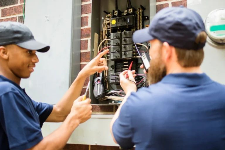 Two workers in navy caps inspecting an open electrical panel outside a building