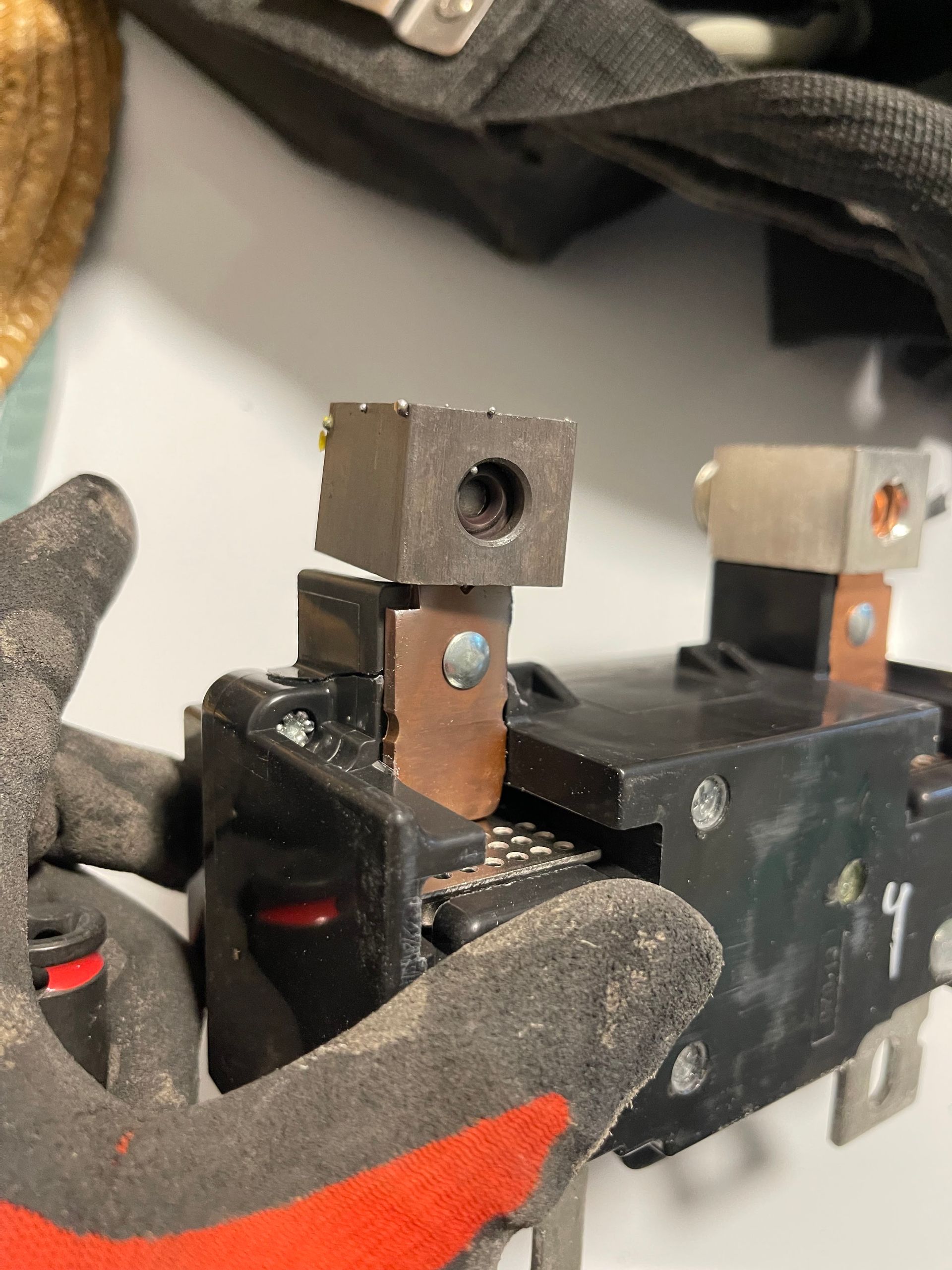 Gloved hand holding a black mechanical part with metal connectors and screws.