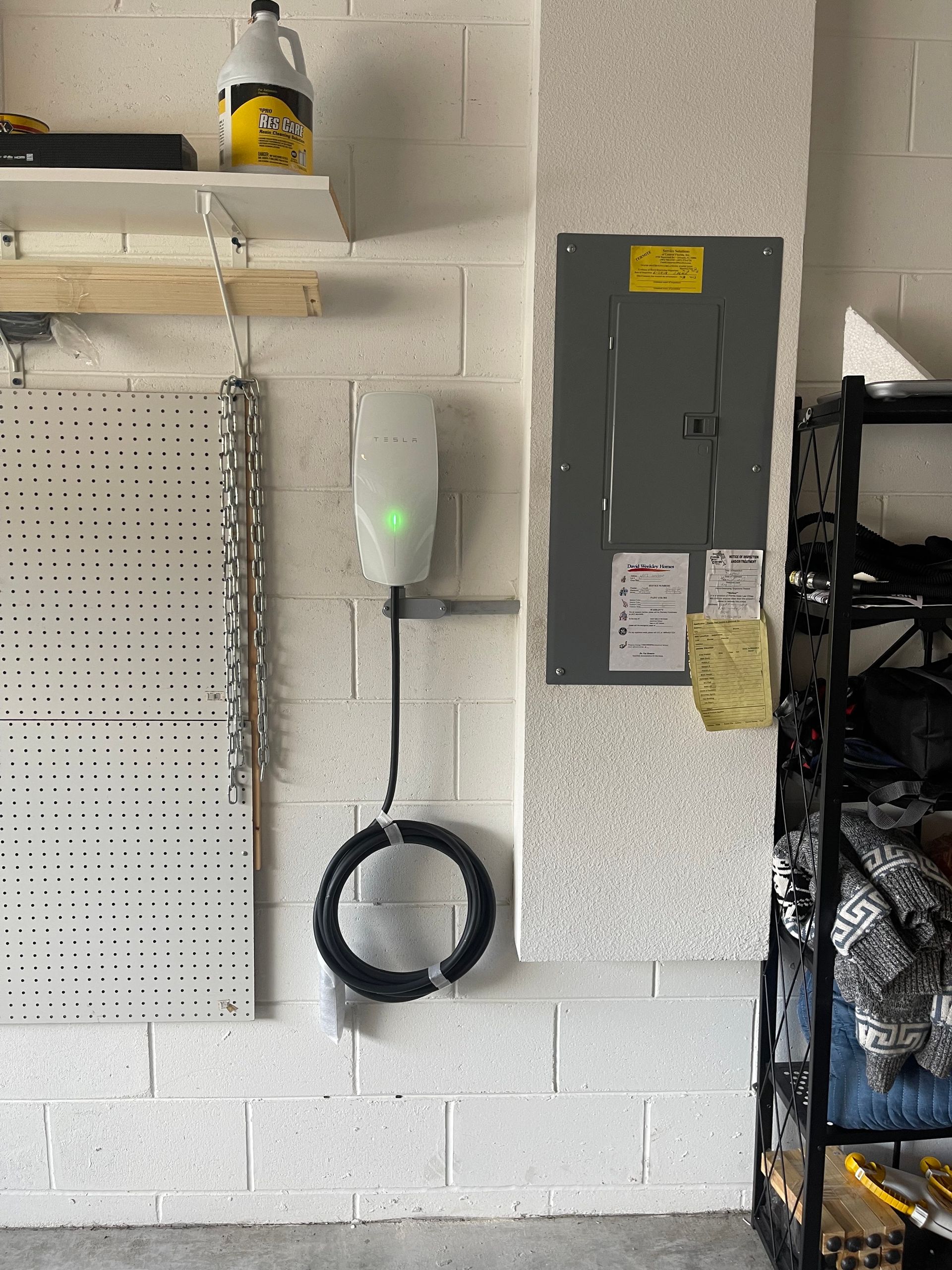 Garage wall with mounted white device, coiled black cable, and electrical panel beside a pegboard.