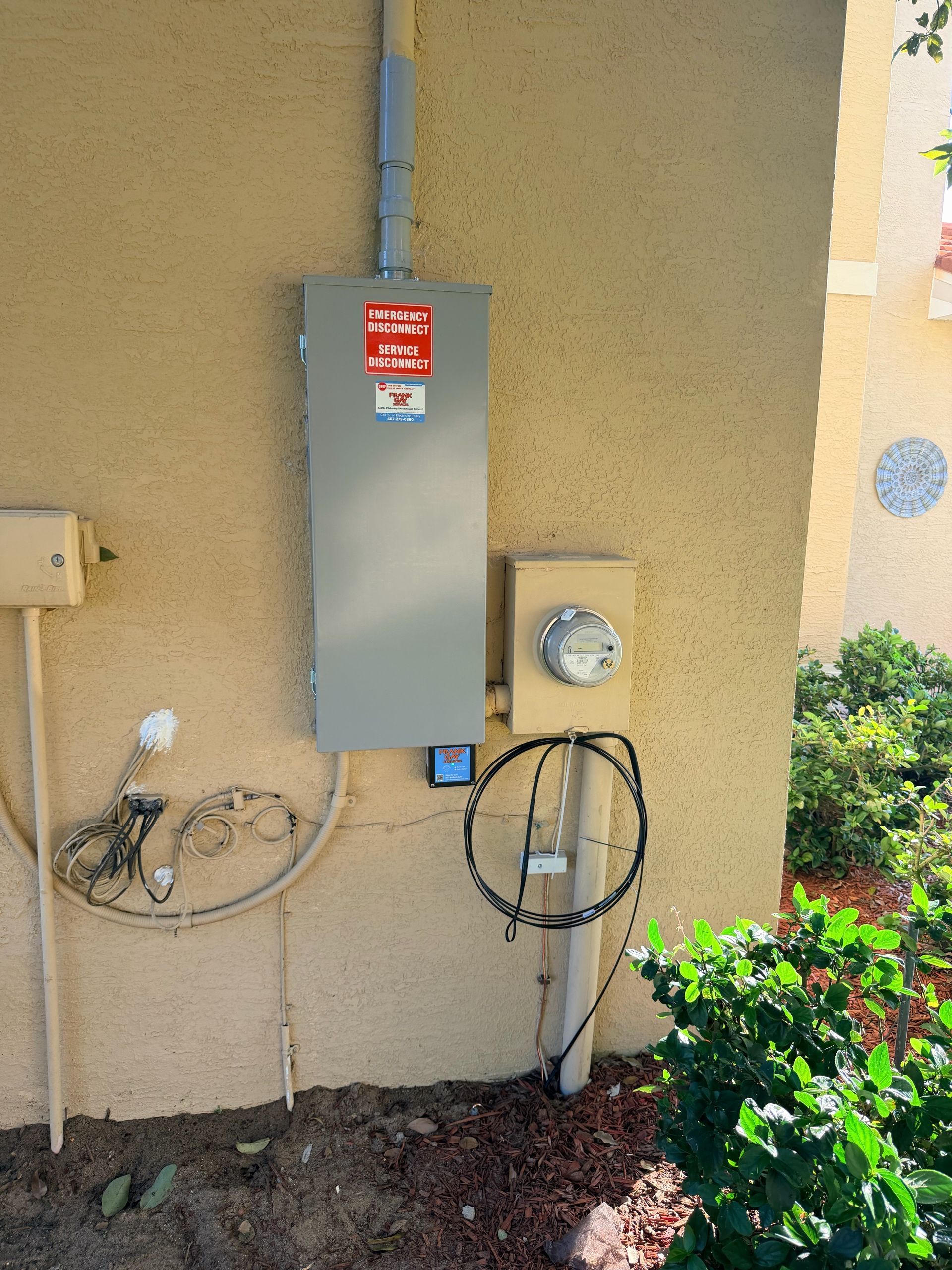 Gray electrical panels and meters mounted on a stucco wall beside plants and dirt.