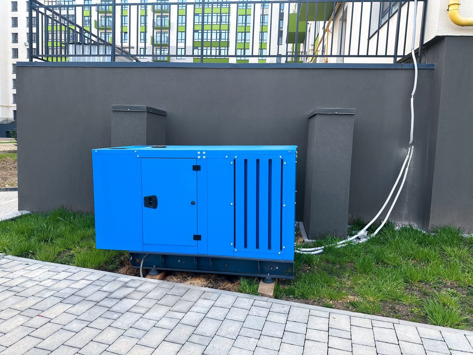 Blue outdoor generator unit beside a gray wall with cables and grass